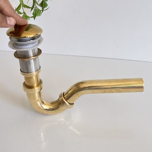 May include: A gold-colored brass drain pipe with a curved elbow and a round, gold-colored drain cover. The drain cover has a white rubber seal.