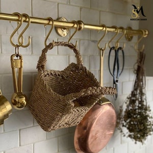 Könnte beinhalten: Eine Küchenleiste aus Messing mit Haken, an denen ein geflochtener Korb, eine Kupferpfanne und eine Gartenschere hängen. Die Leiste ist an einer weißen gekachelten Wand montiert.