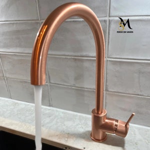 Op de afbeelding: Een koperen keukenkraan met een hoge boog en een hendel. Water stroomt uit de tuit.