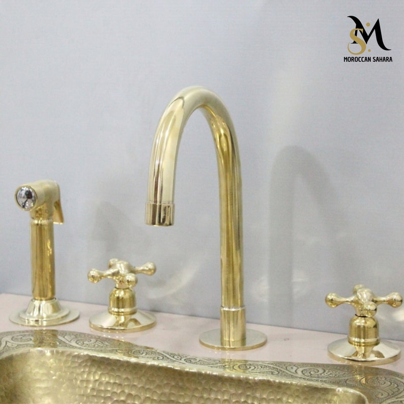 May include: A gold-colored bathroom faucet set with a curved spout, two cross-shaped handles, and a side sprayer. The set is made of brass and has a polished finish. The sink has an ornate hammered design.