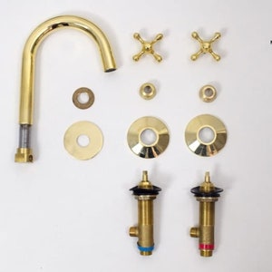 May include: A disassembled gold-colored faucet set, including a curved spout, cross-shaped handles, and various washers and fittings. The components are arranged against a white background, showcasing the Moroccan Sahara logo.