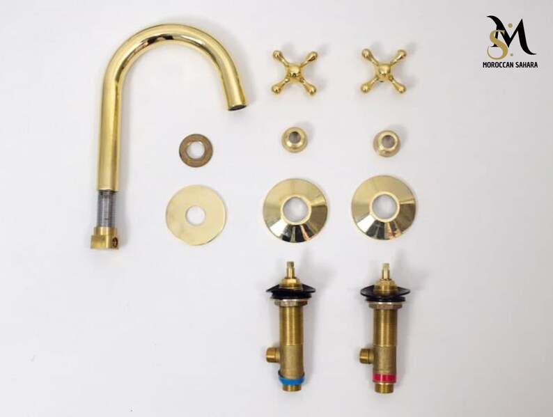 May include: A disassembled gold-colored faucet set, including a curved spout, cross-shaped handles, and various washers and fittings. The components are arranged against a white background, showcasing the Moroccan Sahara logo.