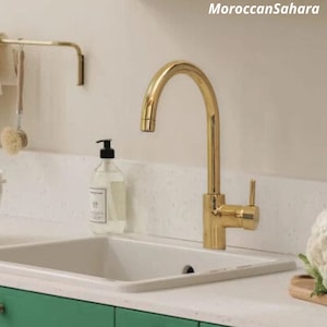 May include: A gold kitchen faucet with a single handle and a high arc spout. The faucet is mounted on a white countertop.