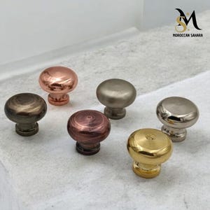 Peut inclure: Six boutons de porte de différentes couleurs, chacun avec un dessus rond et une base ronde plus petite. Les boutons sont en métal et ont une finition polie. Les couleurs sont le cuivre, l'argent, le bronze, l'or et deux nuances de gris.