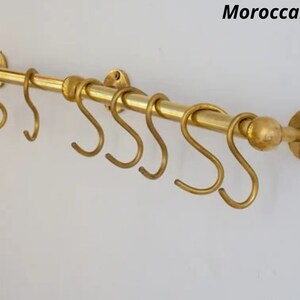 Puede incluir: Un colgador de ollas de metal dorado con seis ganchos. El colgador está montado en una pared y tiene una perilla redonda en el extremo.