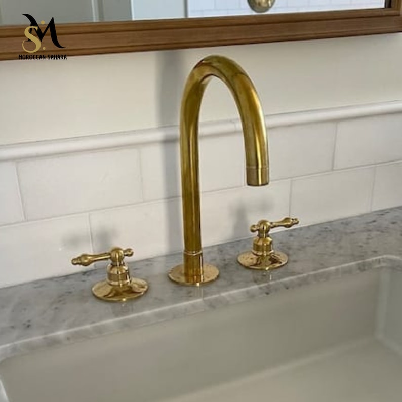 May include: A gold-colored bathroom faucet with a curved spout and two separate handles. The faucet is installed on a marble countertop with a white sink and white tile backsplash. The faucet has a classic design.