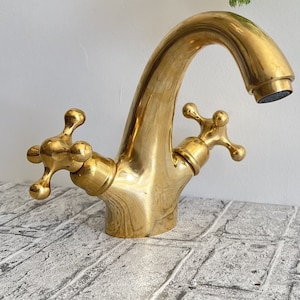 May include: A gold-toned bathroom faucet with two cross-shaped handles. The faucet has a curved spout and a round base.