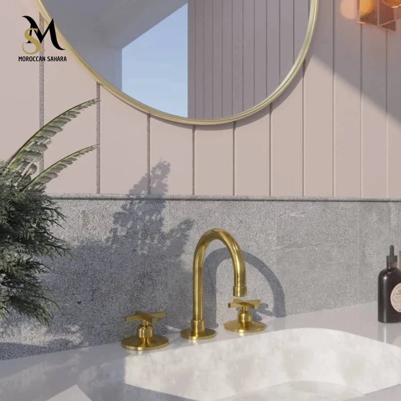 May include: A gold-toned bathroom faucet with cross-shaped handles and a curved spout is mounted on a white countertop. The faucet is set against a backdrop of a round gold-framed mirror and a light pink wall. The text "MOROCCAN SAHARA" is visible.
