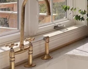 Op de afbeelding: Een gouden keukenkraan met twee handvatten en een hoge uitloop. De kraan is gemonteerd op een wit aanrecht.