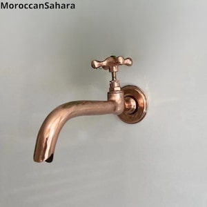 Könnte beinhalten: Ein kupferner Wandwasserhahn mit einem kreuzförmigen Griff. Der Wasserhahn hat einen langen Auslauf und eine runde Basis.