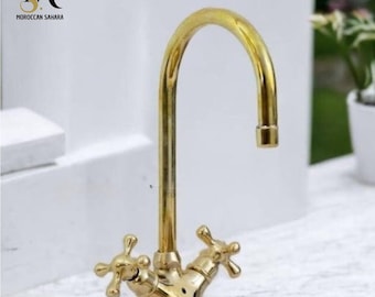 May include: A gold-toned faucet with a high arch spout and two cross-shaped handles. The faucet is mounted on a white marble countertop.