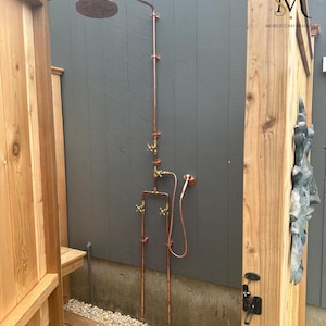 May include: Outdoor shower with copper pipes and brass fixtures. The shower head is round and has a copper finish. The shower is mounted on a wooden wall.