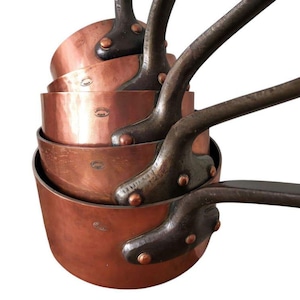 May include: A set of copper saucepans with dark metal handles, stacked together. The copper pots vary in size and are nested. The copper has a reddish-brown hue, and the handles are attached with rivets.