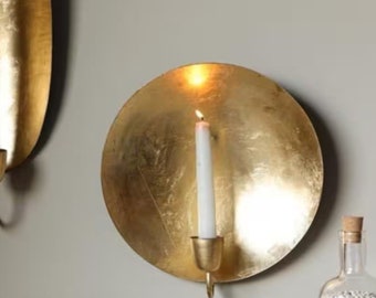 May include: Gold wall-mounted candle holder with a round, textured design. A white candle is lit inside the holder.