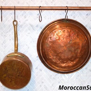 May include: Copper kitchenware hanging on a copper rod with hooks. The kitchenware includes a copper pot and a copper strainer. The rod is mounted on a white tiled wall.