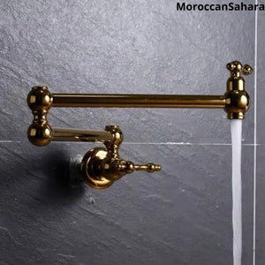 May include: A gold-colored, wall-mounted pot filler faucet with a classic design. The faucet extends horizontally and features a water stream flowing from the spout. The text "MoroccanSahara" is in the upper right corner.
