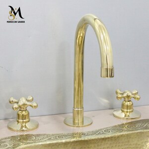 May include: A gold-colored bathroom faucet set with a curved spout and two cross-shaped handles. The faucet is mounted on a light pink surface. The logo "MOROCCAN SAHARA" is visible in the upper left corner.