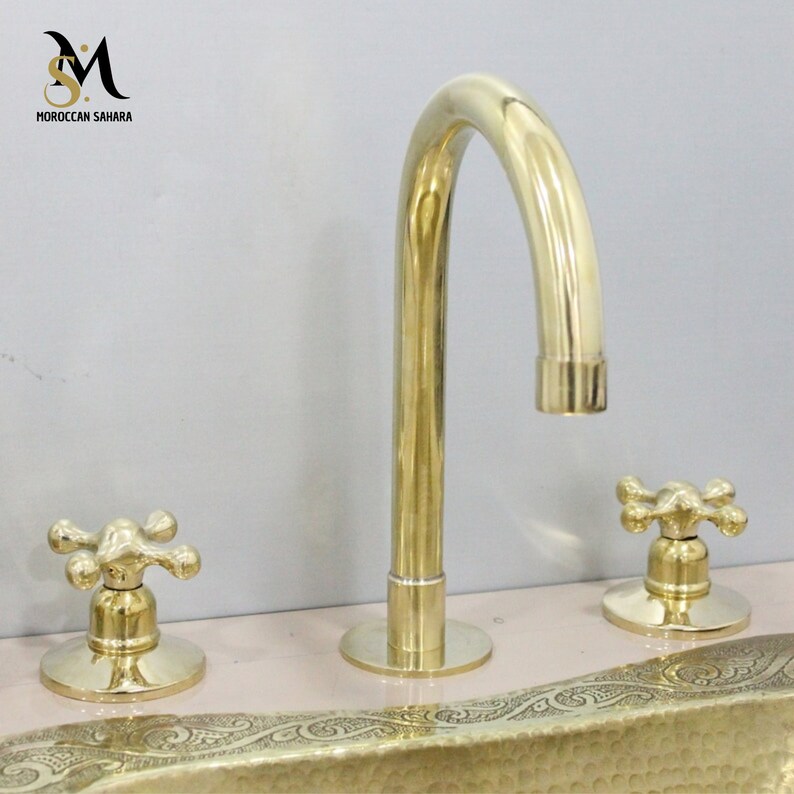 May include: A gold-colored bathroom faucet set with a curved spout and two cross-shaped handles. The faucet is mounted on a light pink surface. The logo "MOROCCAN SAHARA" is visible in the upper left corner.