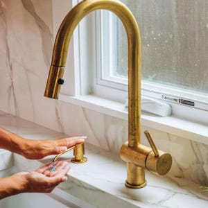 May include: A gold kitchen tap with a single handle and a soap dispenser. The tap is mounted on a white countertop with a marble pattern.