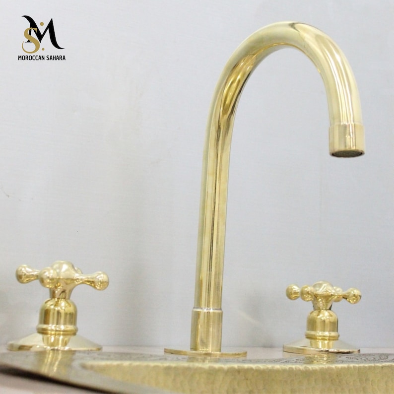 May include: A polished brass faucet with a curved spout and two cross-shaped handles. The faucet is set against a light background. The text "MOROCCAN SAHARA" is visible in the upper left corner.