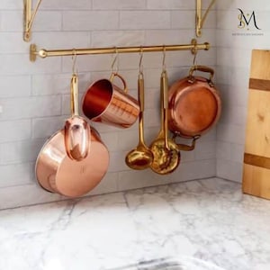 May include: A set of copper kitchen utensils hanging on a brass rack. The utensils include a large copper pot, a copper mug, and two copper ladles. The rack is mounted on a white tiled wall.