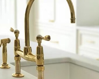 Op de afbeelding: Een gouden keukenkraan met twee aparte uitlopen en een hoog boogontwerp. De kraan heeft een gepolijste afwerking en is gemonteerd op een wit aanrecht.