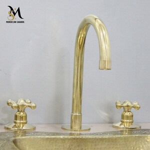 May include: A polished brass faucet with a curved spout and two cross-shaped handles. The faucet is part of a bathroom fixture set. The brand name "MOROCCAN SAHARA" is visible in the upper left corner.