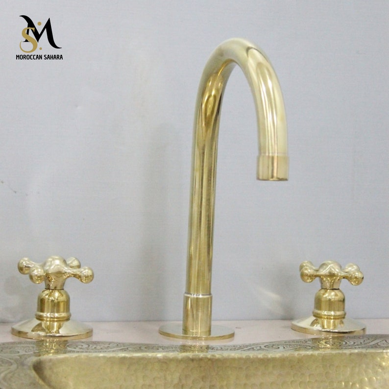 May include: A polished brass faucet with a curved spout and two cross-shaped handles. The faucet is part of a bathroom fixture set. The brand name "MOROCCAN SAHARA" is visible in the upper left corner.