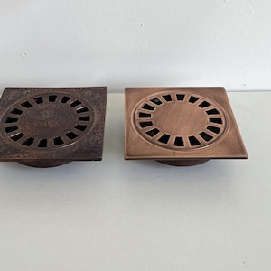 Könnte beinhalten: Zwei quadratische Abflussabdeckungen mit einem runden Design. Die Abflussabdeckungen sind aus kupferfarbenem Metall gefertigt und haben eine gehämmerte Oberfläche. Die Abflussabdeckungen sind beide gleich groß und gleich geformt.
