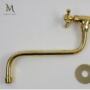 Op de afbeelding: Een gouden messing kraan met een kruisvormige hendel en een gebogen uitloop. De kraan is ontworpen voor wandmontage en bevat een ronde, platte, decoratieve plaat.