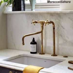 May include: A white porcelain kitchen sink with a brass faucet with two handles. The faucet has a long, curved spout. The sink is on a white marble countertop.