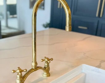 Op de afbeelding: Een gouden keukenkraan met een hoge boog en twee kruisvormige handvatten. De kraan is gemonteerd op een wit aanrecht.
