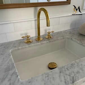 May include: A gold-toned bathroom faucet with a curved spout and two separate handles. The faucet is installed on a white sink with a marble countertop. A decorative drain stopper is visible in the sink.