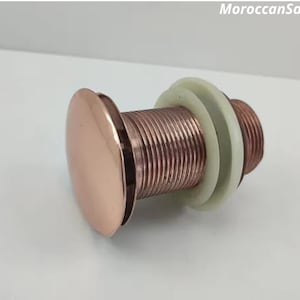 Könnte beinhalten: Ein roségoldener Waschbecken-Ablaufstopfen mit einem Gewindekörper aus Metall und einer runden, glatten Oberseite.