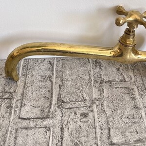 Puede incluir: Un grifo de lat&oacute;n dorado con una manija en forma de cruz. El grifo tiene un ca&ntilde;o largo y curvo y est&aacute; montado en una pared.