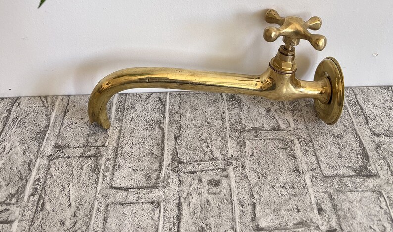 Puede incluir: Un grifo de lat&oacute;n dorado con una manija en forma de cruz. El grifo tiene un ca&ntilde;o largo y curvo y est&aacute; montado en una pared.
