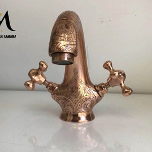 Può includere: Un rubinetto color rame con intricati motivi incisi. Il rubinetto ha due maniglie a forma di croce e un beccuccio curvo. Il testo "MOROCCAN SAHARA" è visibile nell'angolo in alto a sinistra.