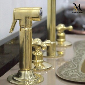 May include: Close-up of a polished brass bidet sprayer with a lever and a round base. Other brass fixtures are in the background, including a faucet and a decorative plate. The logo "MOROCCAN SAHARA" is visible.