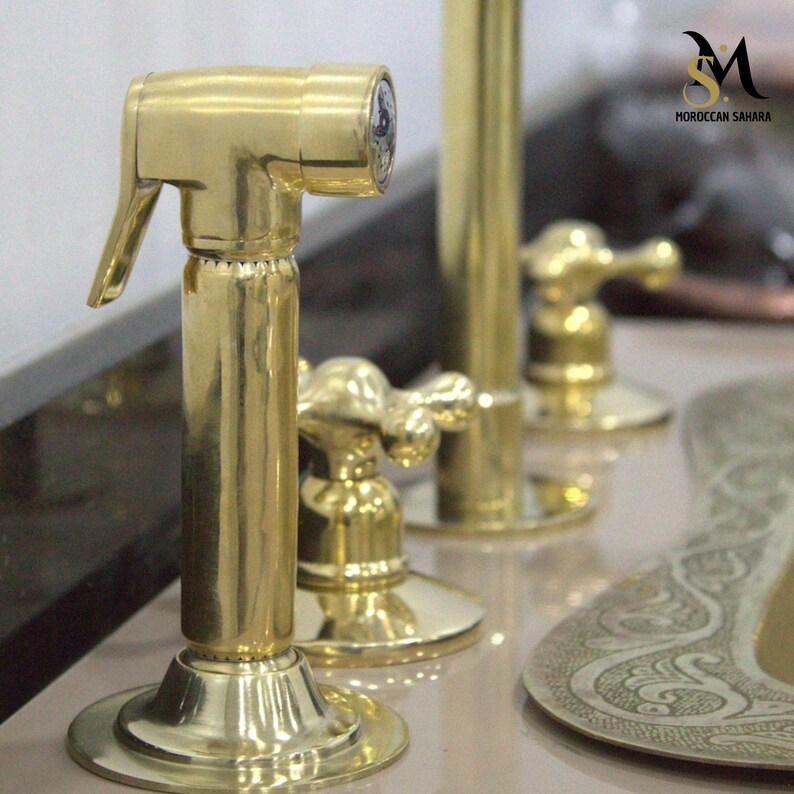 May include: Close-up of a polished brass bidet sprayer with a lever and a round base. Other brass fixtures are in the background, including a faucet and a decorative plate. The logo "MOROCCAN SAHARA" is visible.