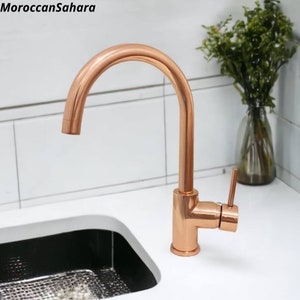 Puede incluir: Un grifo de cocina de cobre cepillado con una sola manija y un caño de arco alto. El grifo está montado en una encimera blanca.