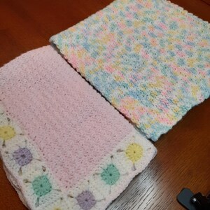 May include: Two crocheted baby blankets. One is pink with a white border and colorful flowers. The other is white with a multi-colored pattern.