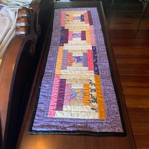 May include: A rectangular, handmade quilt in shades of purple, pink, orange, and cream. The quilt features a log cabin design with various patterned fabrics, including floral and butterfly motifs. The quilt is placed on a dark wooden surface.