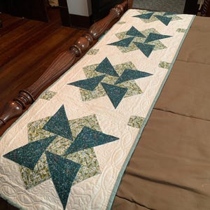 May include: A quilted bed runner featuring a repeating star pattern. The stars are composed of teal and floral print triangles set against a cream background. The runner is edged with a teal border and has a quilted design.