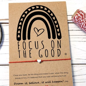 May include: A red string bracelet with a silver heart charm and a card that says "Focus on the good." The card has a black and white rainbow with hearts on it. The card also says "Dream it, believe, it will happen!" and "Handcrafted with love in USA."