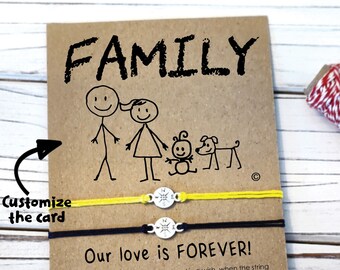 May include: A card with a hand-drawn illustration of a family with two parents, a child, and a dog. The card is decorated with two friendship bracelets with compass charms. The card reads "FAMILY" and "Our love is FOREVER!" with a message about making a wish when the string breaks.