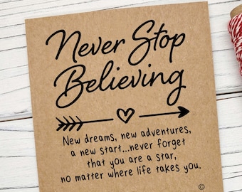 May include: A brown card with the text "Never Stop Believing" and a compass charm bracelet. The card has a quote about new dreams and adventures. The bracelet is navy blue with a silver compass charm.