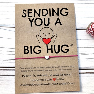 May include: A pink friendship bracelet with a white heart charm and a card that says "Sending you a big hug". The card also includes the text "Close your eyes, tie this string and make a wish, when the string breaks in two, it's believed that your wish will become true! Dream it, believe, it will happen! Handcrafted with in USA MakeaWishXO.com MAWXO.com SparkleXO.com"