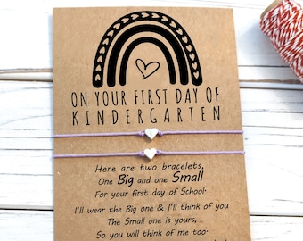 May include: A card featuring a rainbow design with the text "On your first day of Kindergarten". The card includes two bracelets with silver heart charms and a message about wearing one bracelet and thinking of the person who gave it to you.