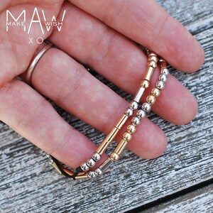 Peut inclure: Deux bracelets avec des perles dorées et argentées sur une ficelle noire. Les bracelets sont empilés l'un sur l'autre. Le texte "MAKE A WISH XO" est visible en arrière-plan.
