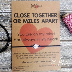 May include: A brown card with the text "Close Together or Miles Apart" and a red heart. The card is attached to a black string bracelet with a silver compass charm. The text "You are on my mind and always in my heart!" is also on the card. The card also includes the text "May your wish come true!" and the website address MakeaWishXO.com.